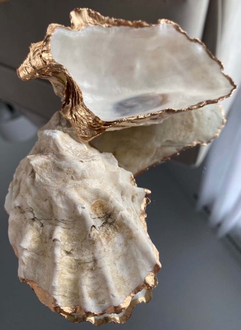 A decorative oyster shell ring dish with a glossy finish and gold leaf trim, displayed as a bridal party gift with pearl accents.