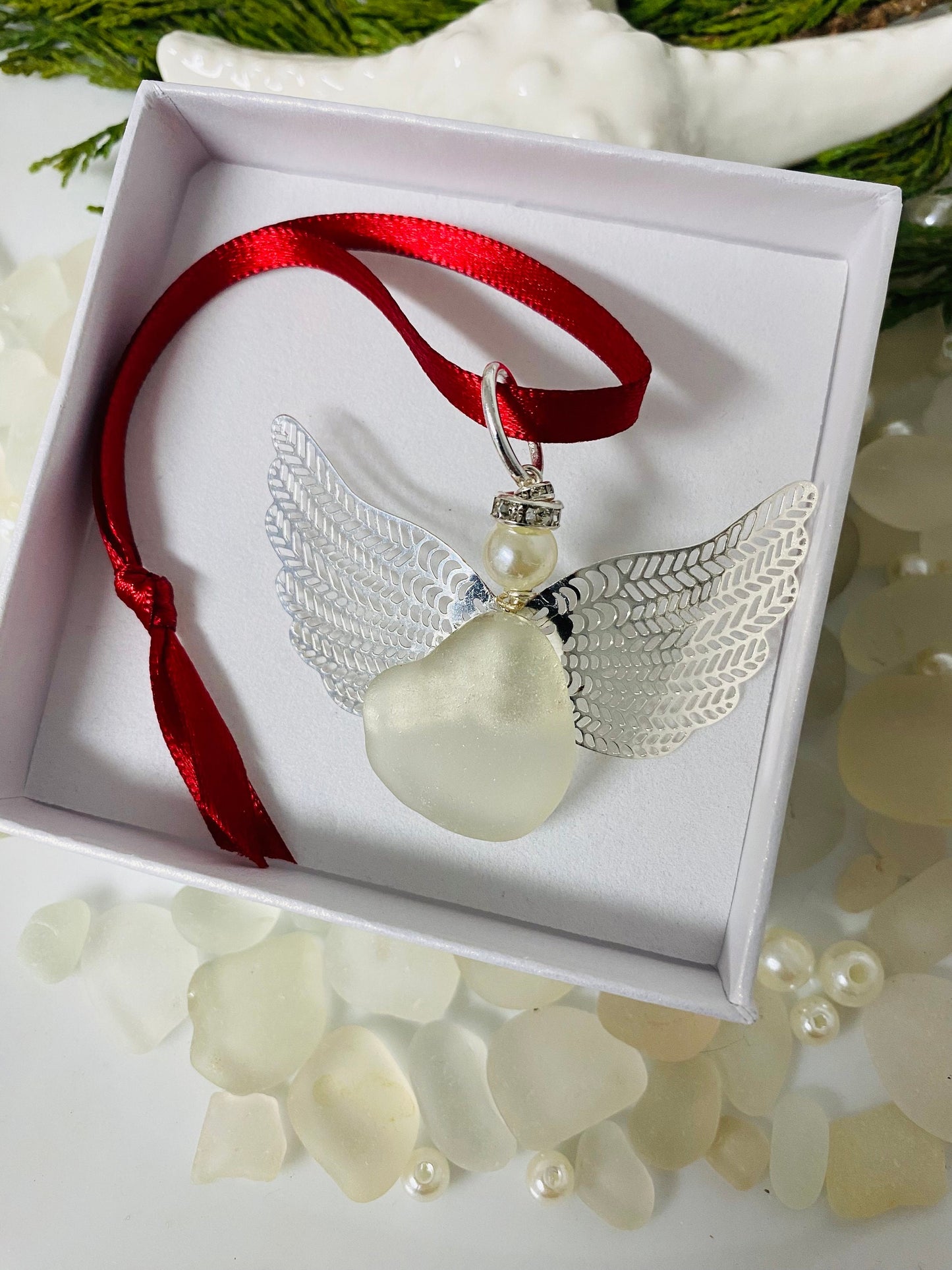 A handcrafted sea glass angel Christmas ornament with a red ribbon, displayed in a white jewelry box.