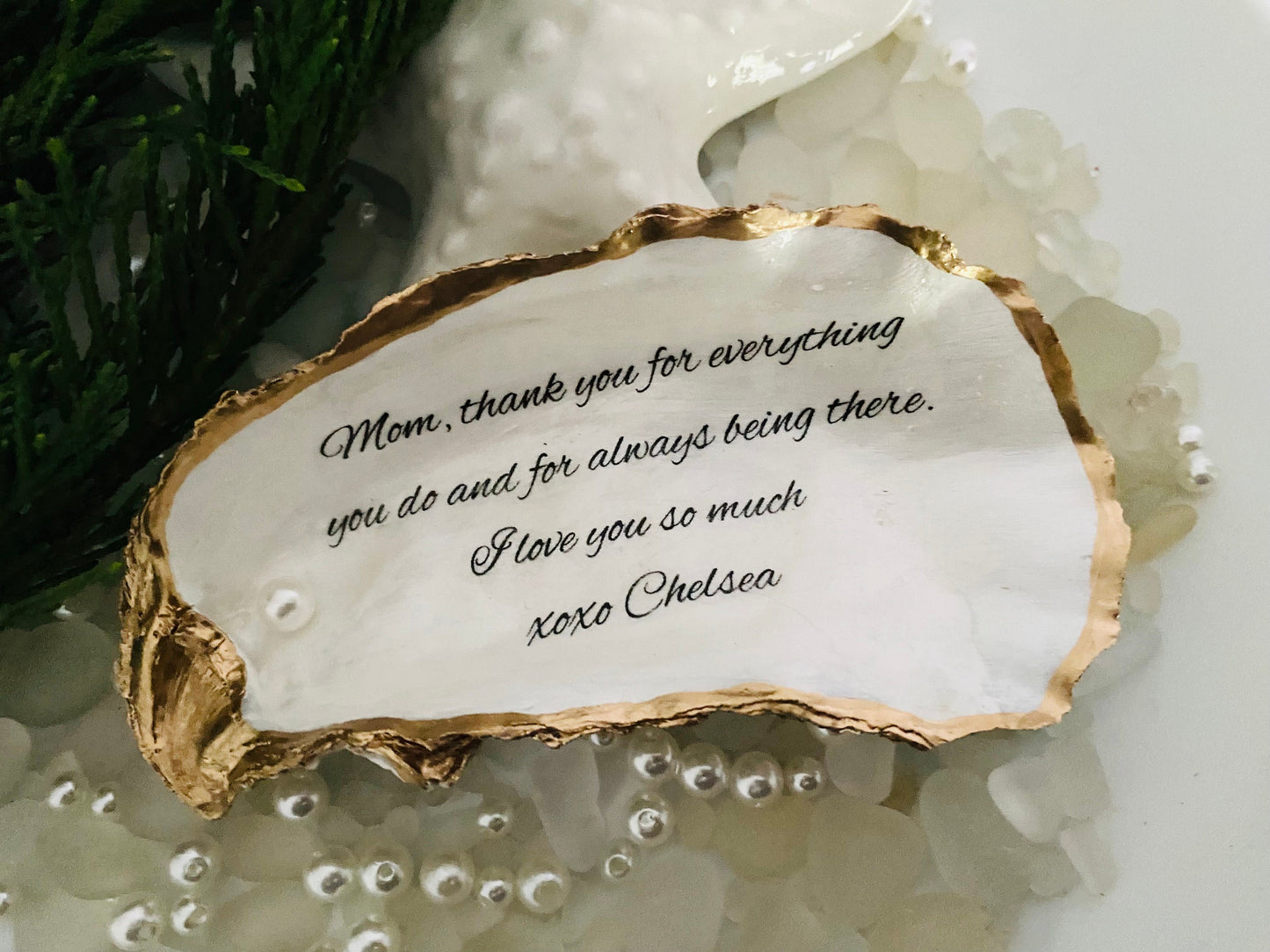 A personalized oyster shell jewelry dish with a message written on it, placed on a white surface surrounded by decorative pearls.