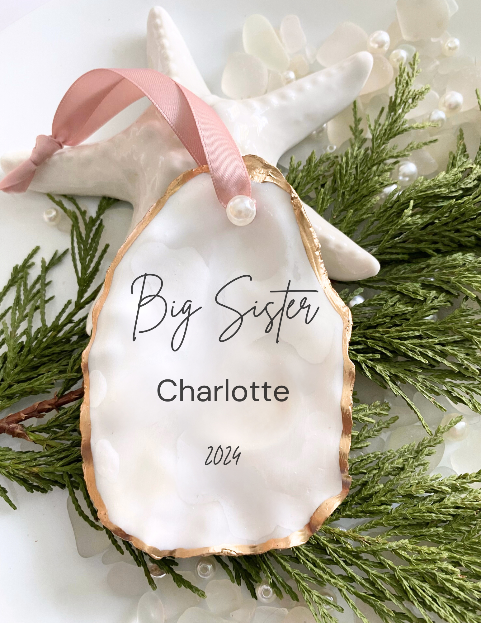 Personalized big sister oyster shell ornament with Charlotte in script, pearl finish, gold trim, and pink ribbon displayed in a white gift box
