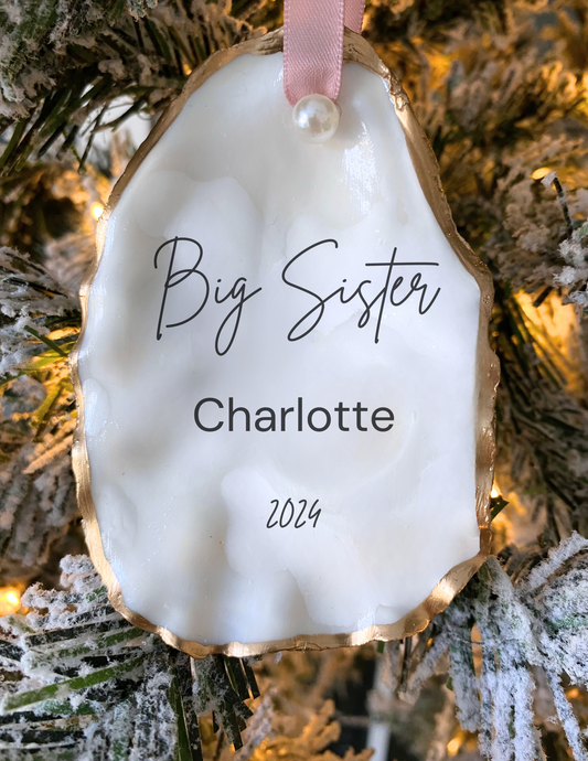 A personalized oyster shell Christmas ornament with 'Big Sister' written on it, along with a name ('Charlotte') and a date ('2024'). The ornament is white with gold gilding and is hanging from a tree.