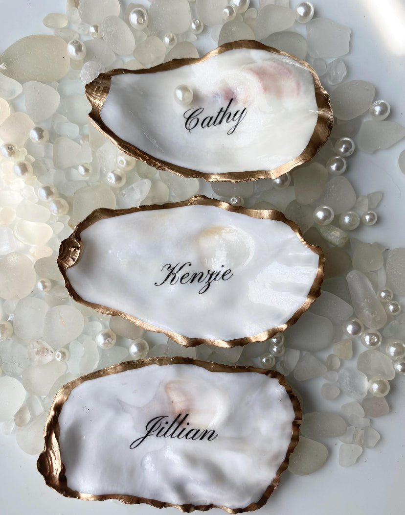 Three oyster shells with gold rims and names engraved on a pearl-studded background