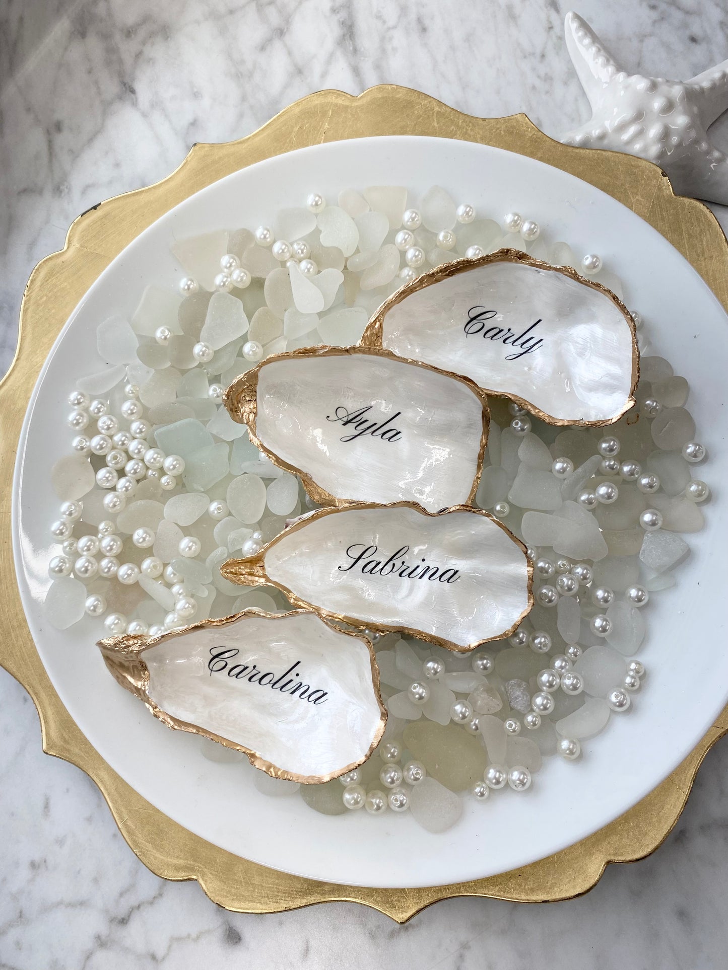 Oyster place card shells with names engraved on them, surrounded by pearls on a marble surface.