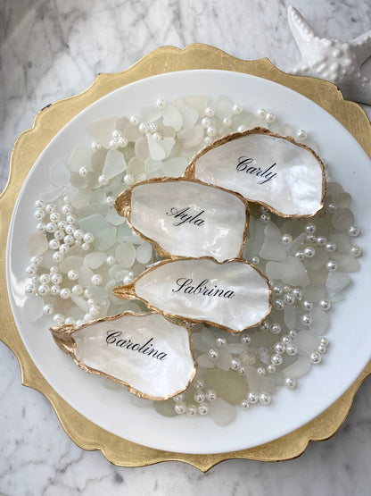 Oyster place card shells with names engraved on them, surrounded by pearls on a marble surface.