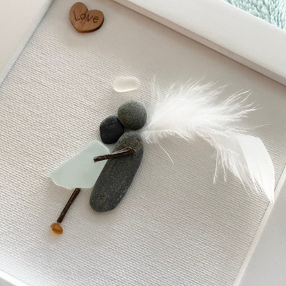 Sea Glass Memorial Framed Art Angel made with pebble embracing a human body with sea glass. Angel has a feather wing, pebble body and halo with a wooden love heart wall art or Desk Top Decor