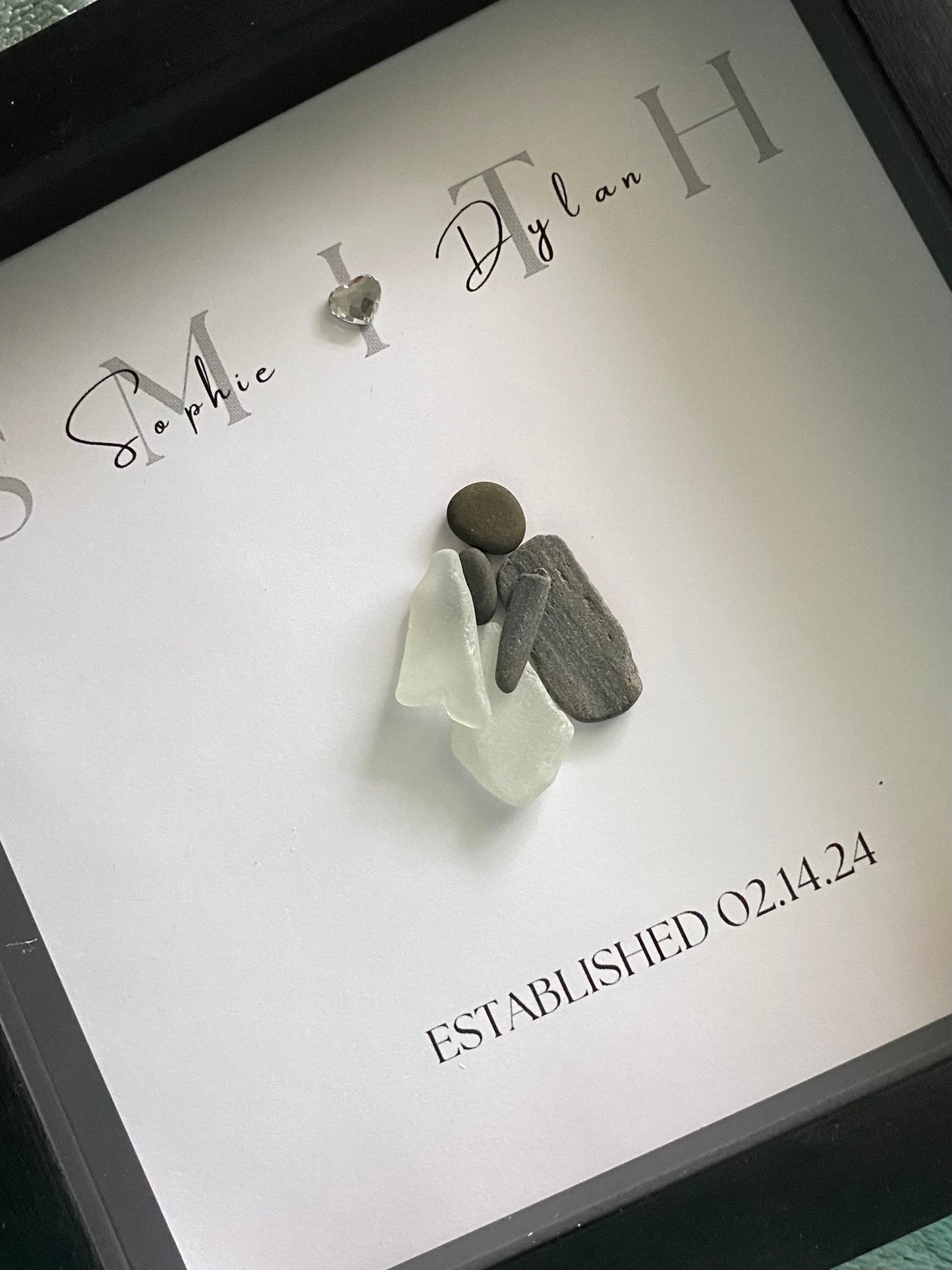 A framed personalized sea glass art piece with the names 'Sophie & Ryan' and a date 'September 7, 2022' written on it, displayed in a black frame.