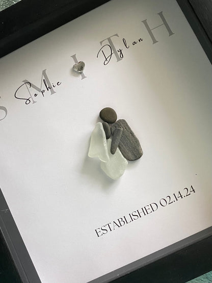 A framed personalized sea glass art piece with the names 'Sophie & Ryan' and a date 'September 7, 2022' written on it, displayed in a black frame.