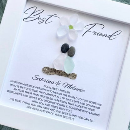 two sea glass friends with pebble heads sitting together on sand with their names and a quote below in a white frame