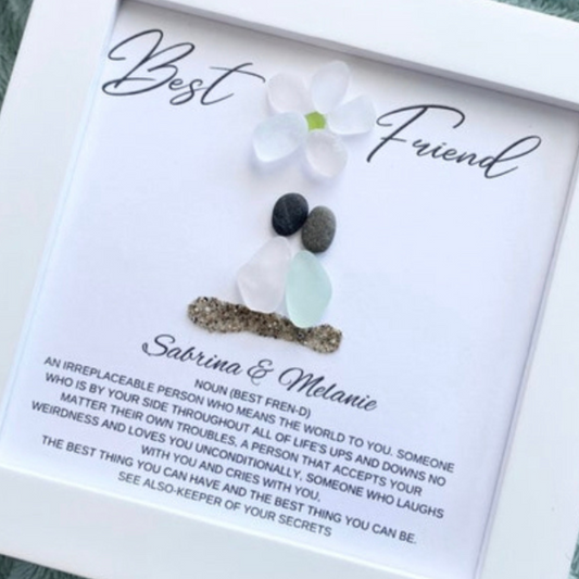 two sea glass friends with pebble heads sitting together on sand with their names and a quote below in a white frame