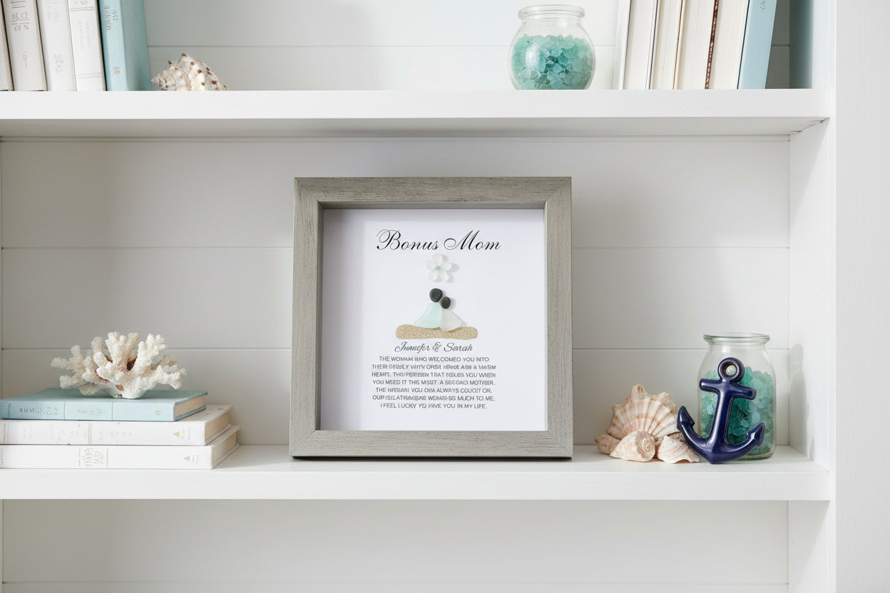 A personalized gift frame with a title 'BONUS MOM' at the top, featuring a sea glass figure and pebble art, placed on a soft surface.