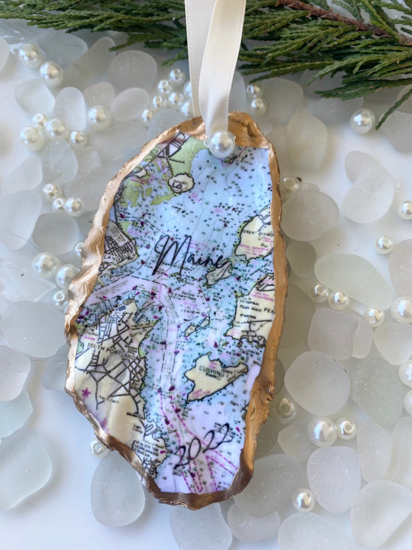 A custom Casco Bay Maine map oyster shell Christmas ornament with the word 'Maine' written on it, personalized with a name and date, displayed on a white background with pearls and greenery.