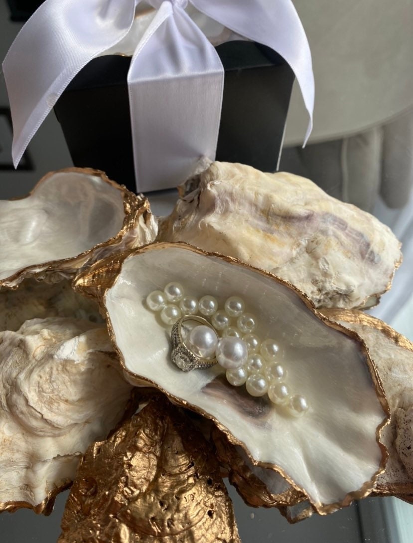 A decorative oyster shell ring dish with a glossy finish and gold leaf trim, displayed as a bridal party gift with pearl accents.
