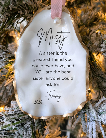 A personalized Christmas ornament oyster shell design, featuring a quote about being a sister, along with greenery and pearls embellishments.