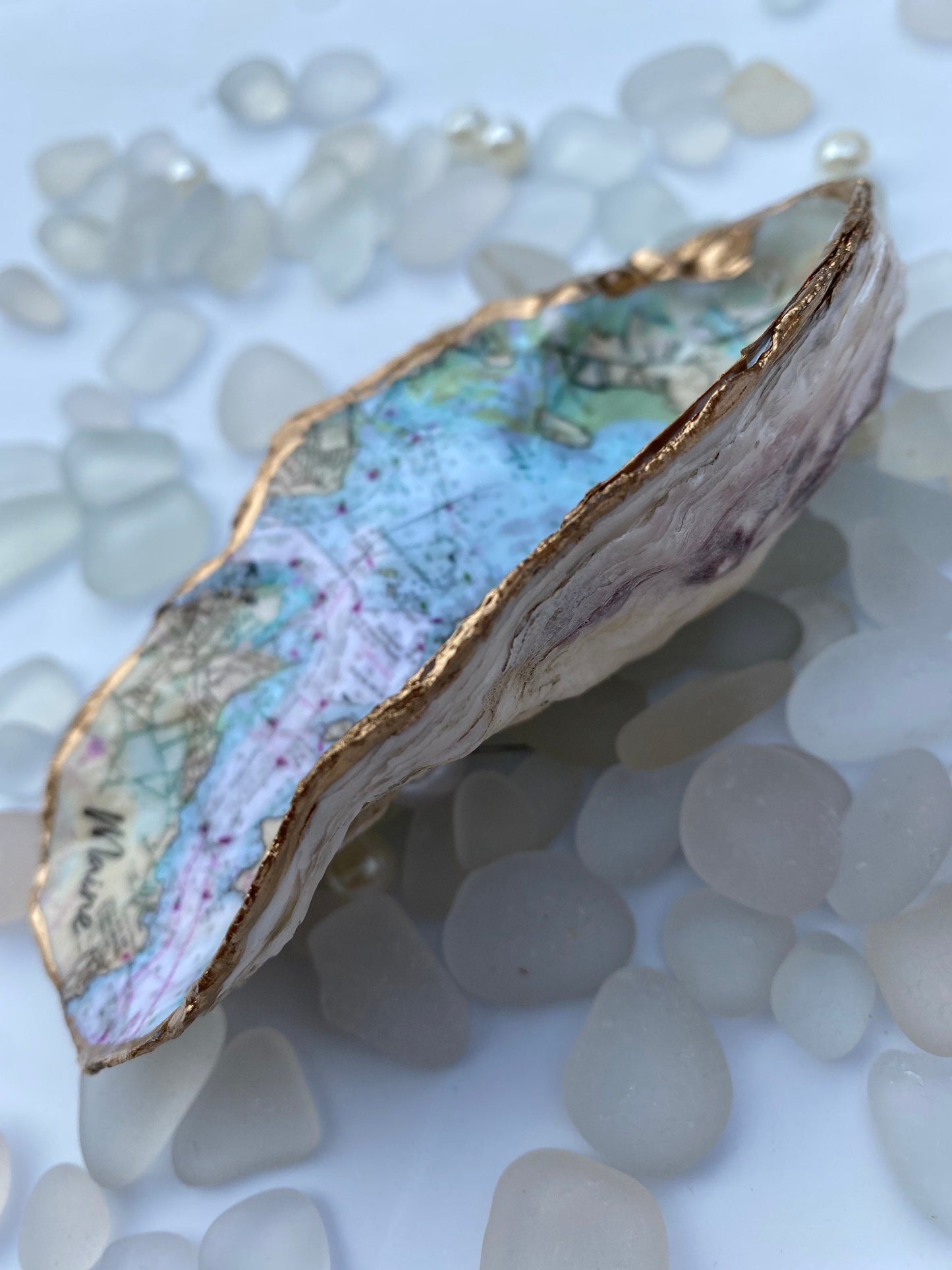 A custom map oyster dish featuring a hand-painted design of the state of Maine, with inlays of pearls and a ring, displayed on a white surface with additional pearls scattered around.