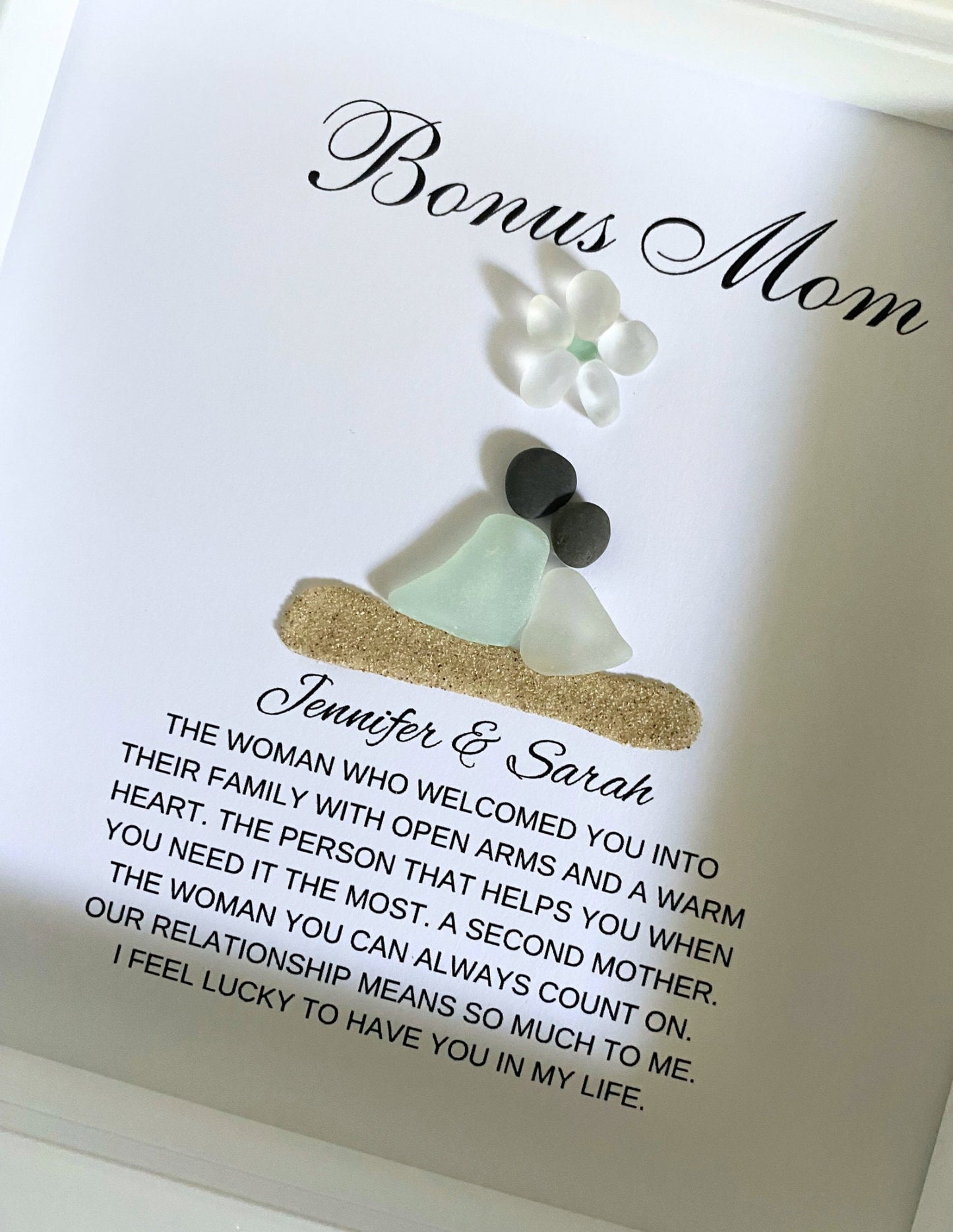A personalized gift frame with a title 'BONUS MOM' at the top, featuring a sea glass figure and pebble art, placed on a soft surface.