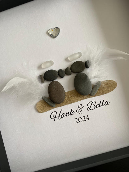 A personalized dog memorial gift featuring a sea glass and pebble art with the names 'Hank & Bella' and a date '2024' inscribed on a piece of paper within a gray frame.
