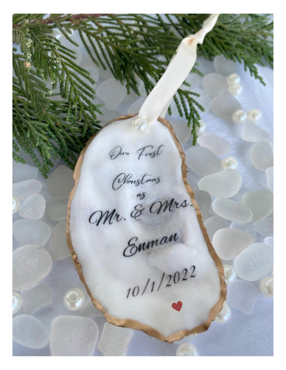 A personalized oyster shell Christmas ornament with the text 'Mr & Mrs' and a date '10/1/2023' on it, hanging from a branch with pearl-like beads and greenery in the background.