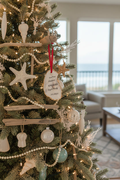 A personalized engagement ornament made of oyster shell with gold gilded edges, displaying a message that reads 'Our First Christmas Engaged', with a date '3.2.2022' below the text. The ornament is hanging from a red ribbon, placed against a backdrop of pine needles.
