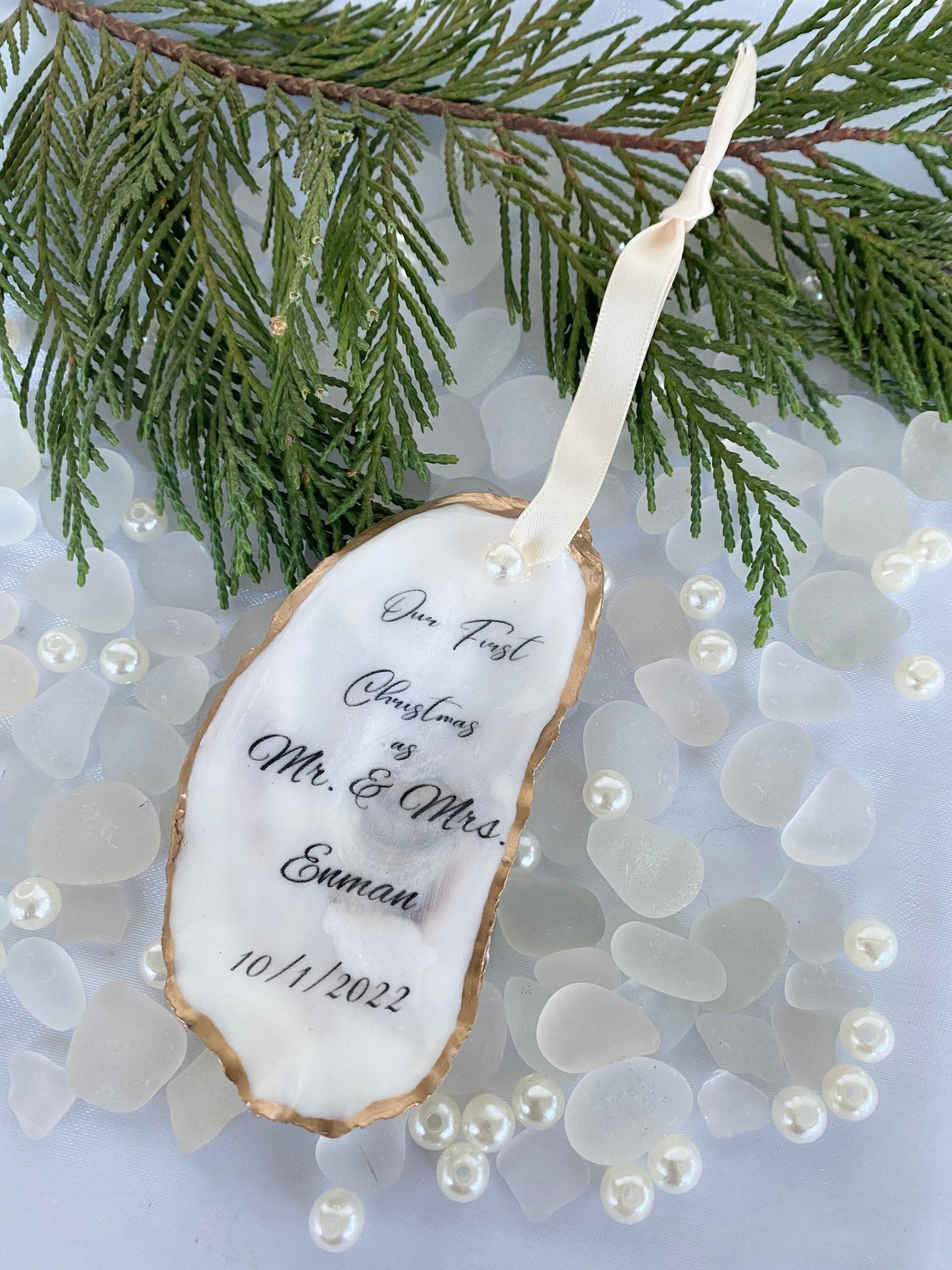 A personalized wedding Christmas ornament made of oyster shell with the names 'Mr & Mrs' and a date '11/27/2024'  on it, placed on a branch with pine needles and decorative pearls around.