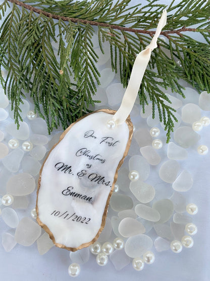 A personalized wedding Christmas ornament made of oyster shell with the names 'Mr & Mrs' and a date '11/27/2024'  on it, placed on a branch with pine needles and decorative pearls around.