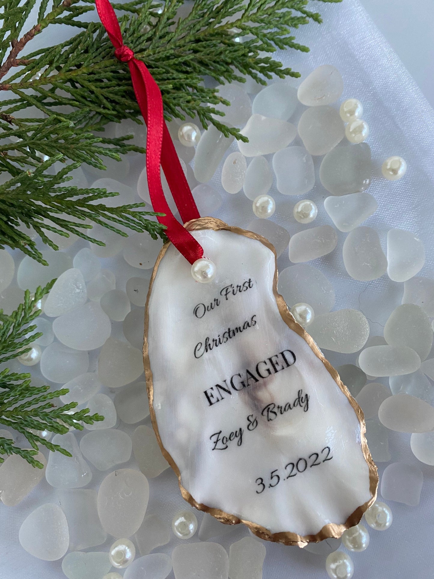 A personalized engagement ornament made of oyster shell with gold gilded edges, displaying a message that reads 'Our First Christmas Engaged', with a date '3.2.2022' below the text. The ornament is hanging from a red ribbon, placed against a backdrop of pine needles.