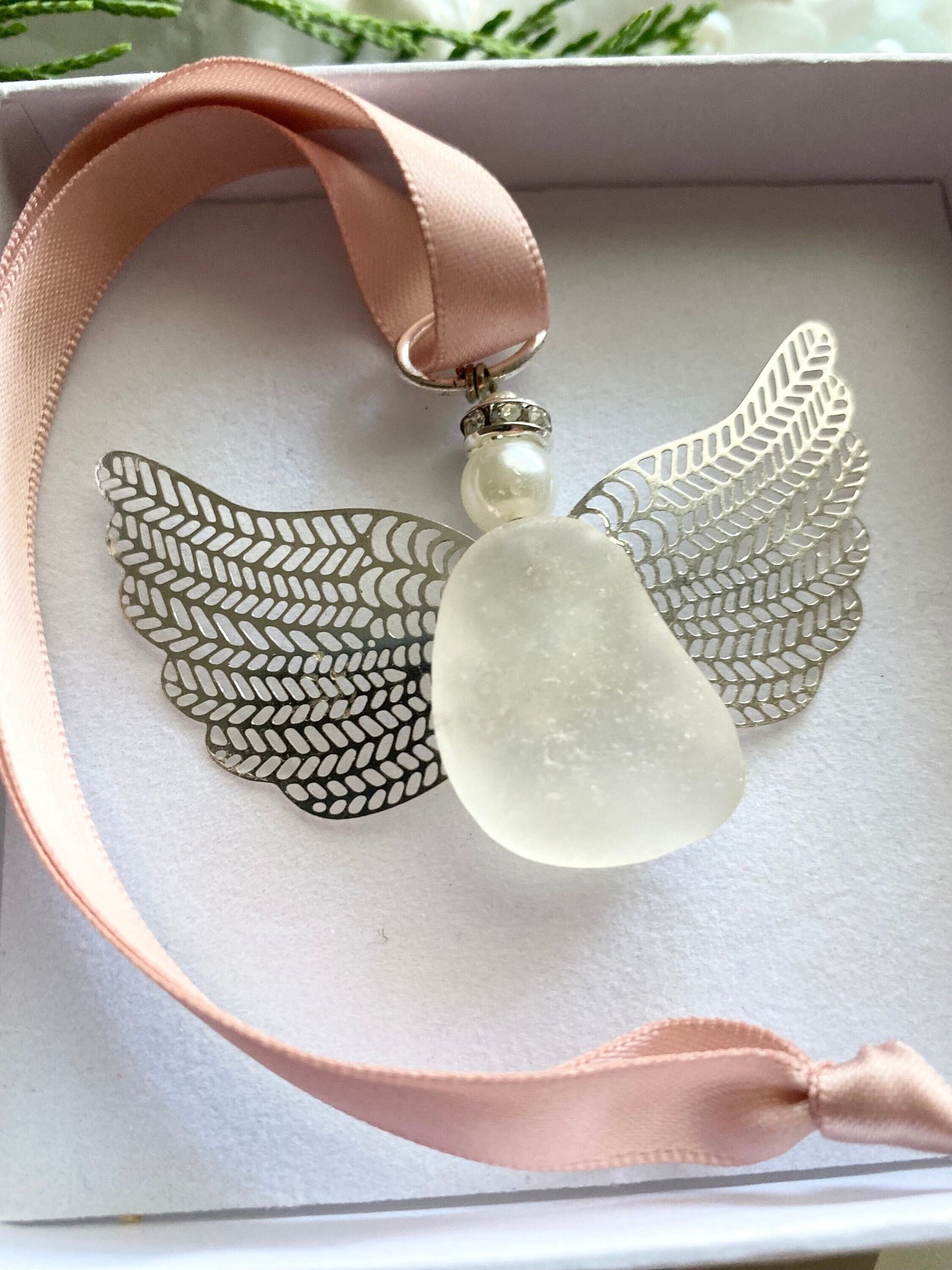 Sea glass angel ornament with a whitish sea glass body, silver metal wings, a rose gold satin ribbon hanger, and a cream pearl-like head inside of a white gift box