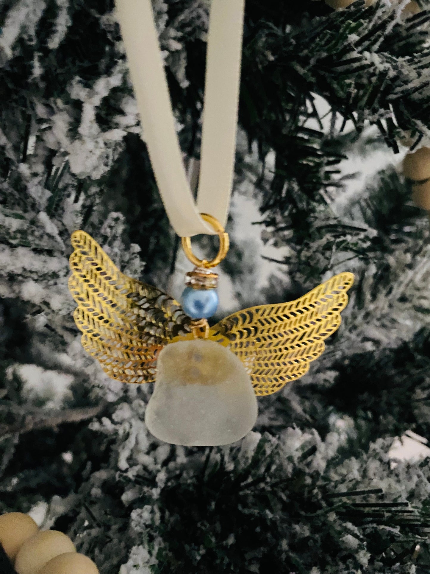 Sea glass angel ornament with a whitish sea glass body, gold metal wings, an ivory satin ribbon hanger, and a blue pearl-like head hanging on a Christmas tree