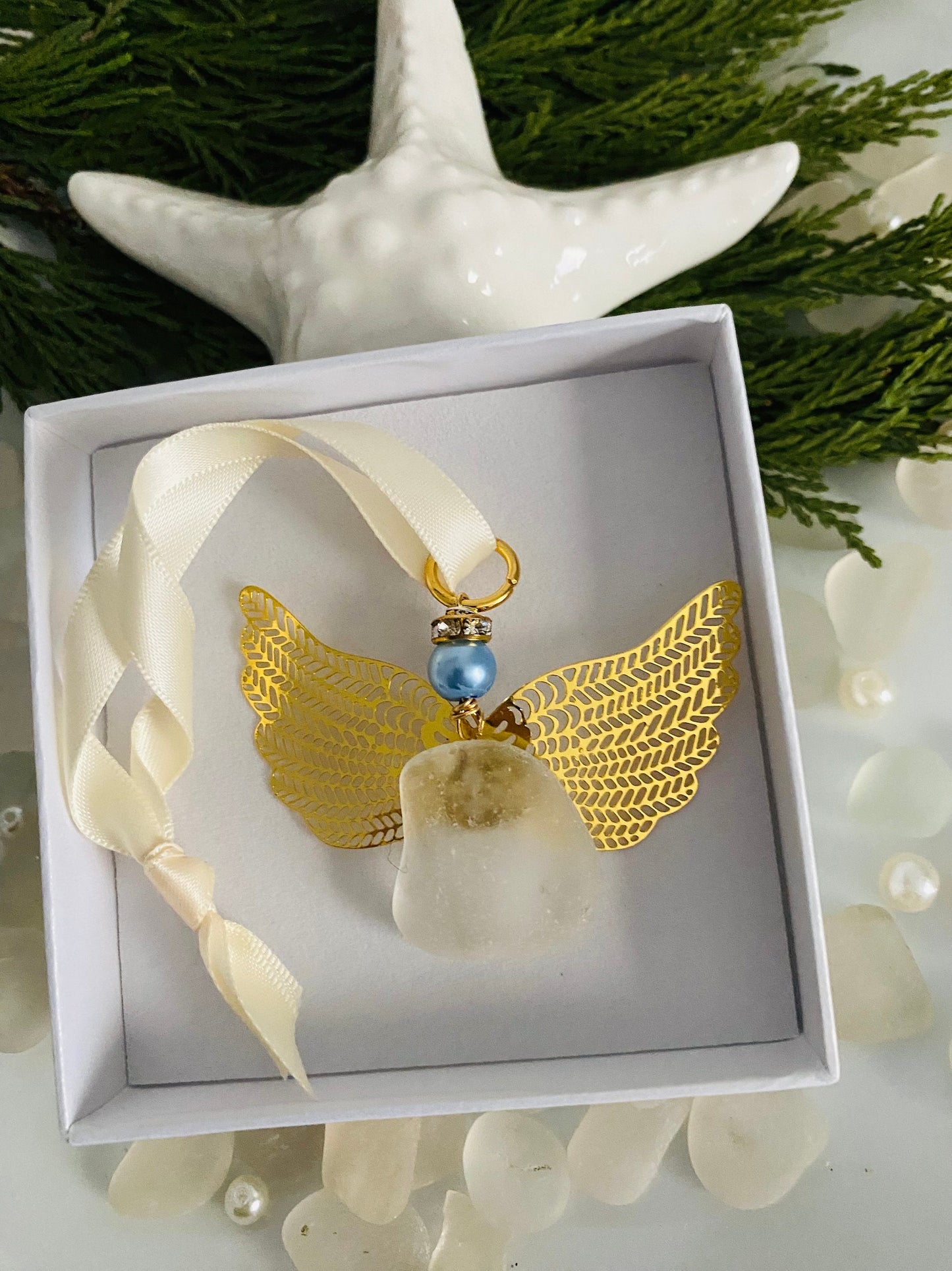 Sea glass angel ornament with a whitish sea glass body, gold metal wings, an ivory satin ribbon hanger, and a blue pearl-like head in a white gift box