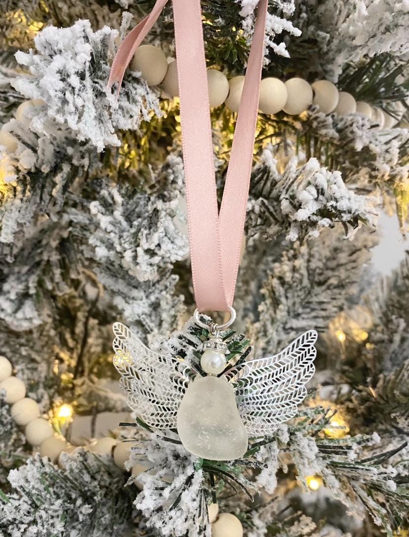 Sea glass angel ornament with a whitish sea glass body, silver metal wings, a rose gold satin ribbon hanger, and a cream pearl-like head hanging on a Christmas tree