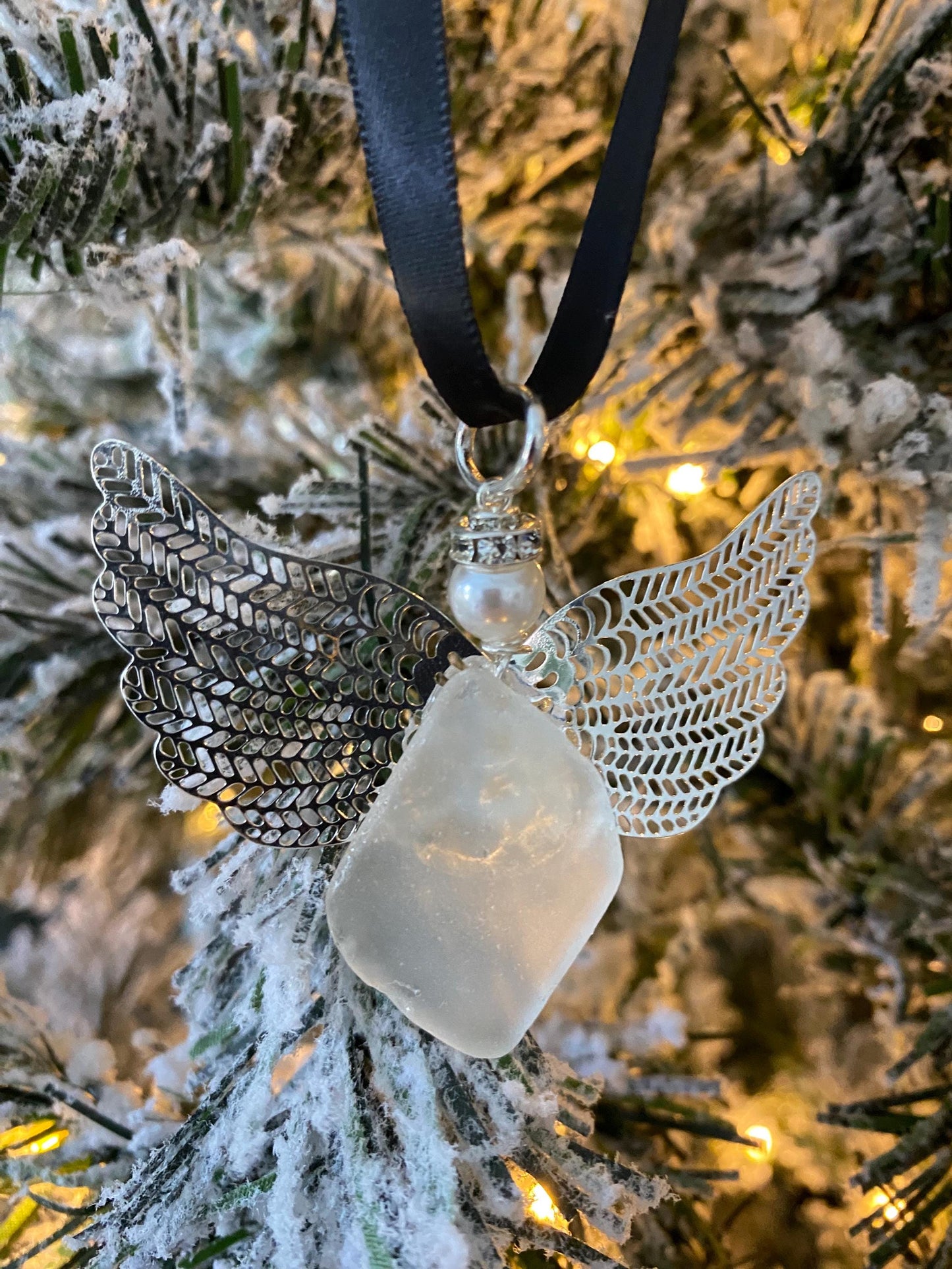 Sea glass angel ornament with a whitish sea glass body, silver metal wings, a black satin ribbon hanger, and a cream pearl-like head hanging on a Christmas tree