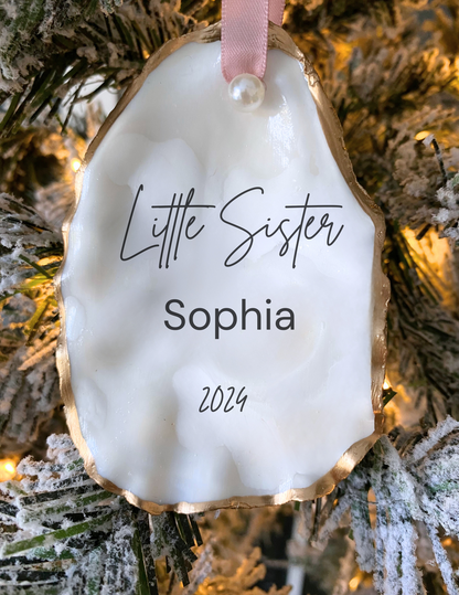 Personalized little sister oyster shell ornament with Sophia in script, pearl finish, gold trim, and pink ribbon displayed in a white gift box