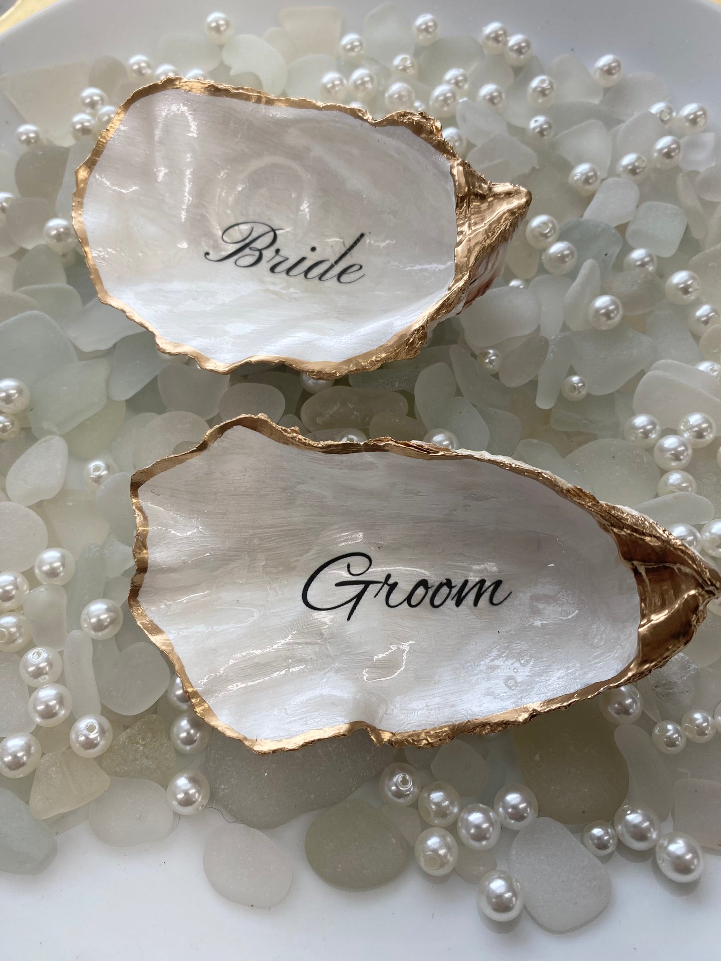 Two oyster shell place cards with 'Bride' and 'Groom' engraved on them, surrounded by pearls.