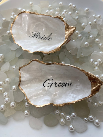 Two oyster shell place cards with 'Bride' and 'Groom' engraved on them, surrounded by pearls.
