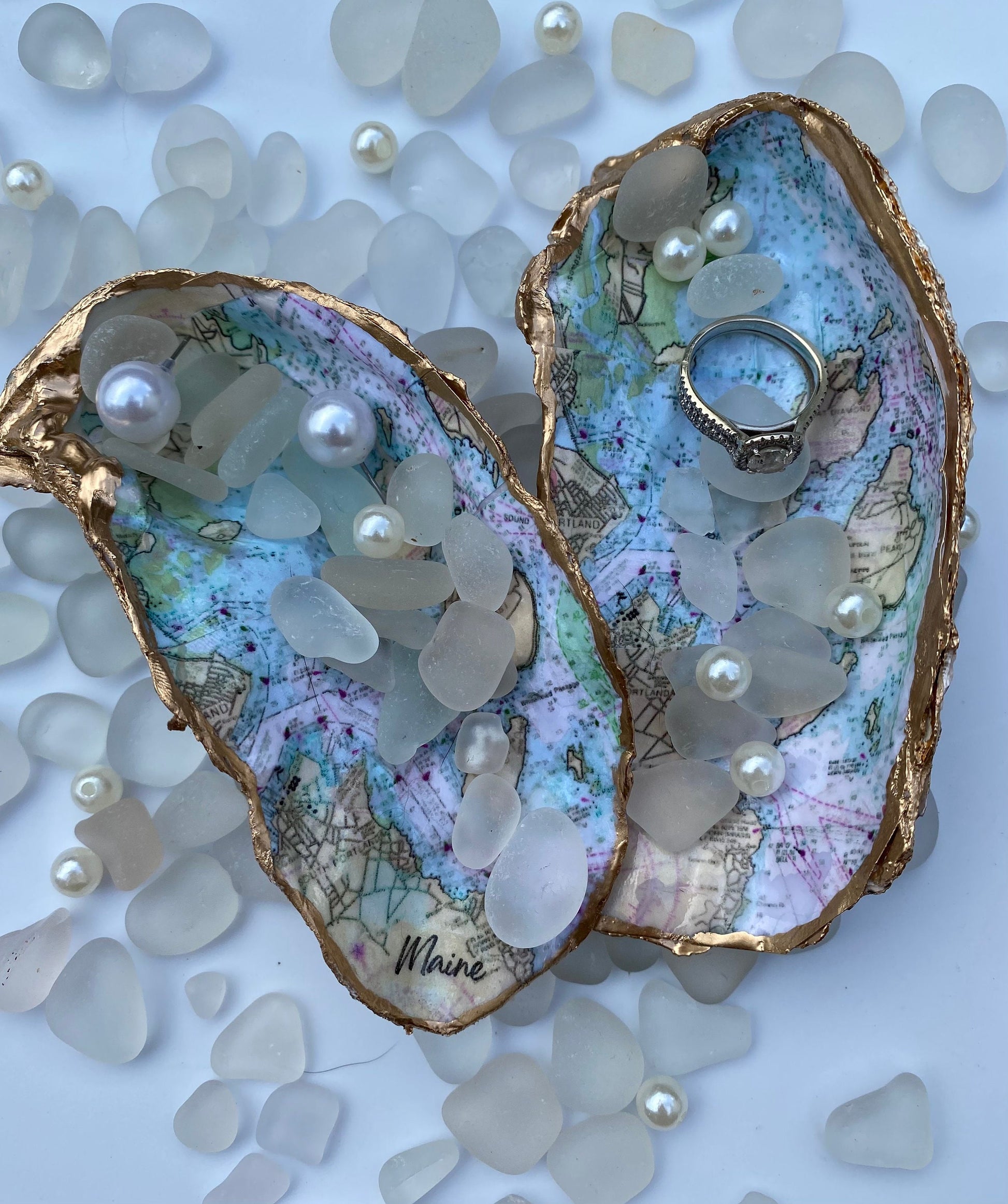 A custom map oyster dish featuring a hand-painted design of the state of Maine, with inlays of pearls and a ring, displayed on a white surface with additional pearls scattered around.