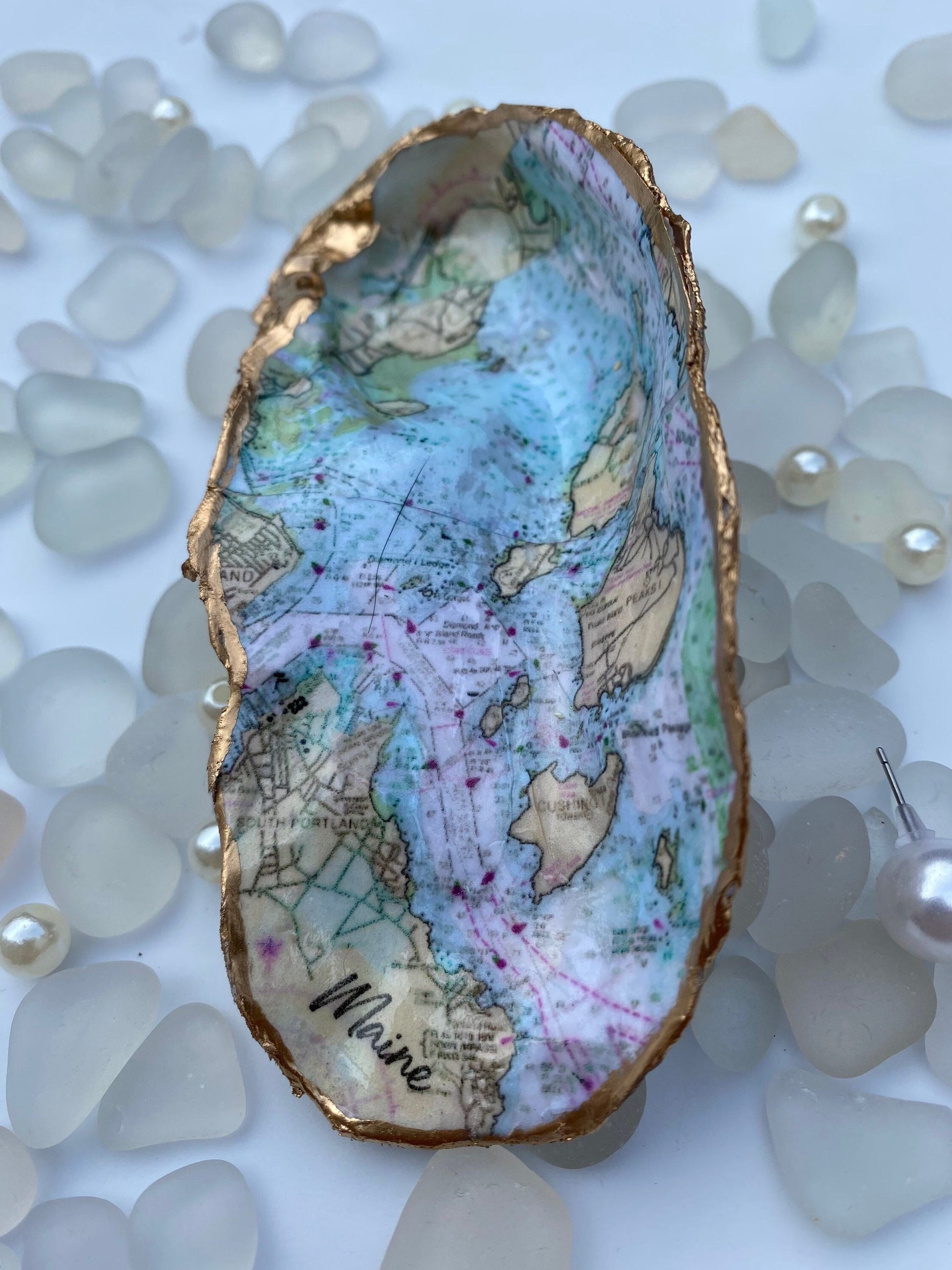 A custom map oyster dish featuring a hand-painted design of the state of Maine, with inlays of pearls and a ring, displayed on a white surface with additional pearls scattered around.