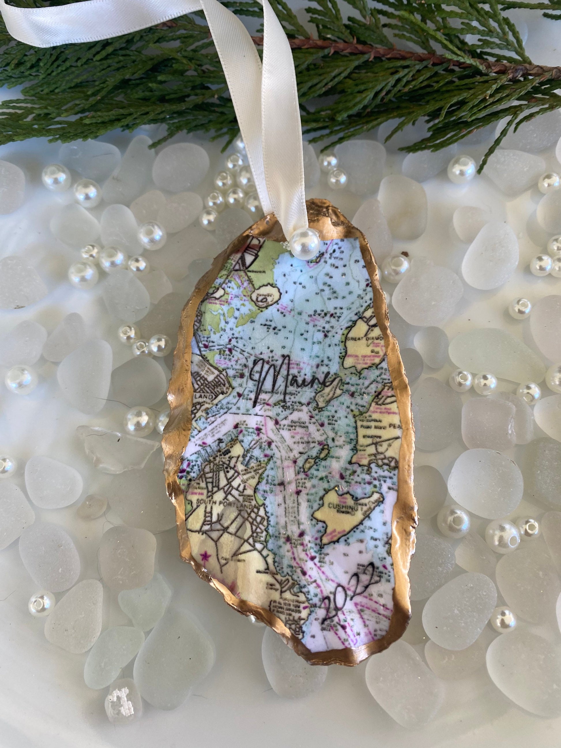 A custom Casco Bay Maine map oyster shell Christmas ornament with the word 'Maine' written on it, personalized with a name and date, displayed on a white background with pearls and greenery.