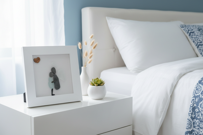 White nightstand with framed artwork and small plant next to a bed with white bedding and decorative pillows.
