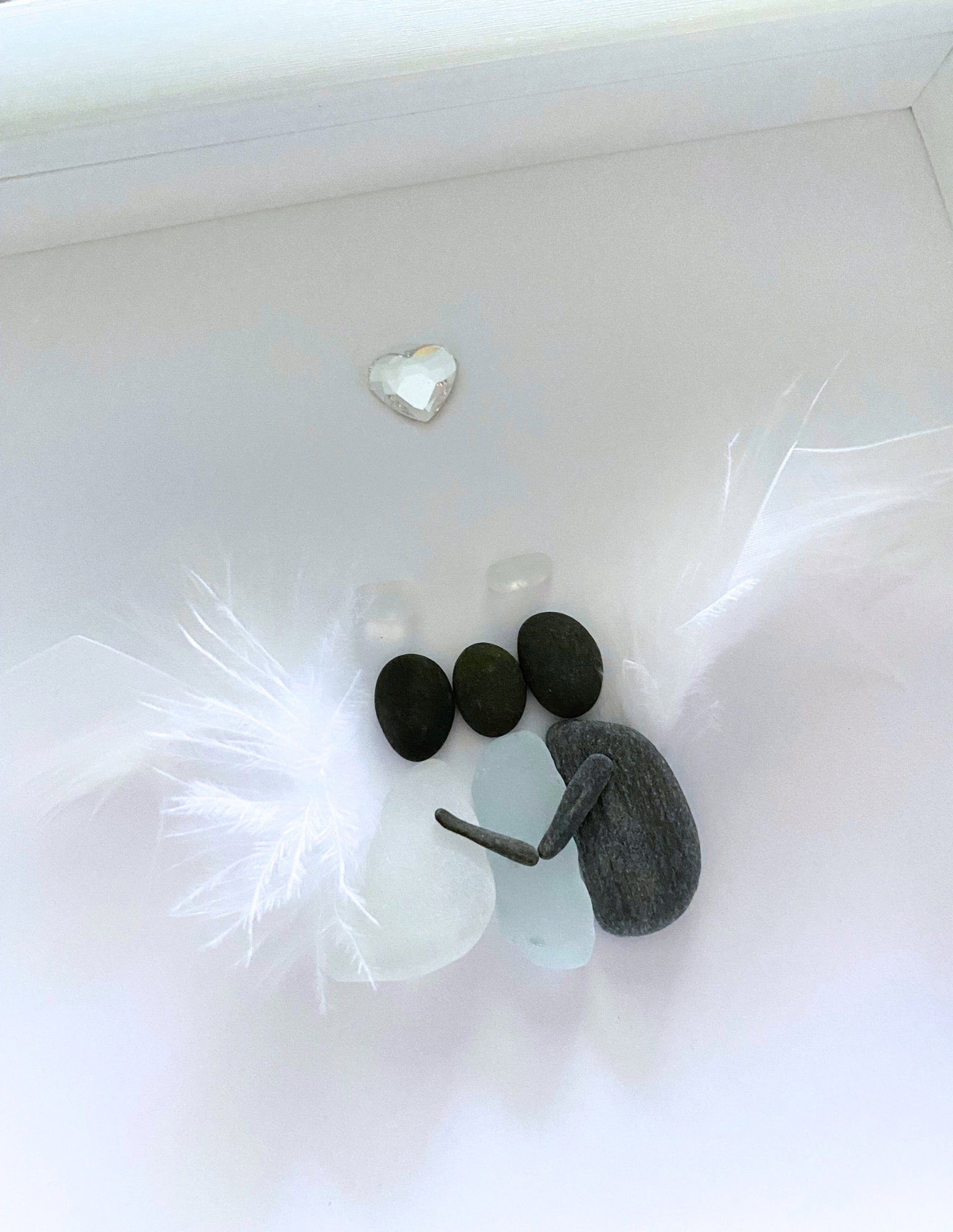 A memorial gift featuring a sea glass art piece with a feather wings hugging a sea glass human, displayed on a white surface.