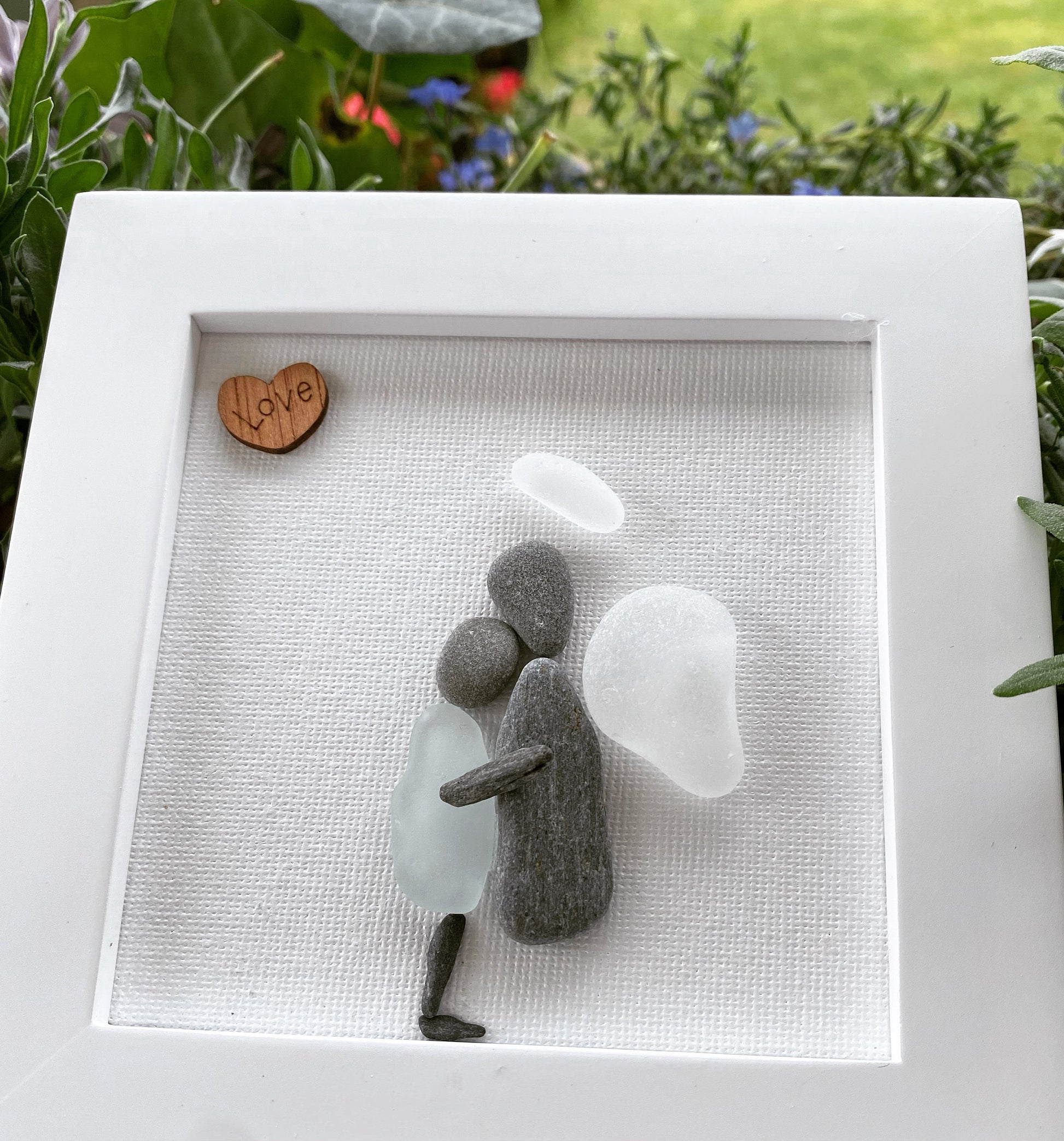 A framed sea glass art piece with a white frame, featuring a picture of a mother embracing her child angel with a wooden heart.
