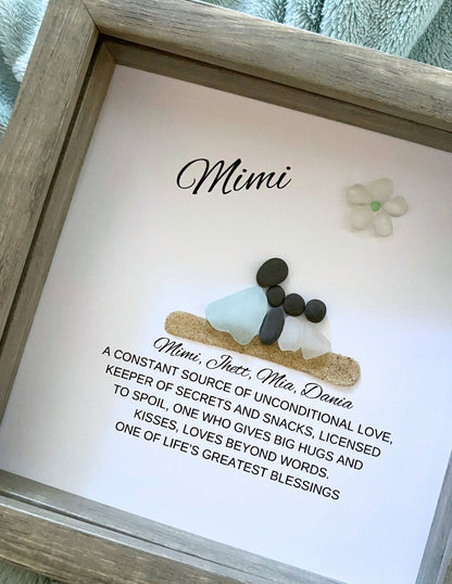A personalized sea glass and pebble art piece with the inscription 'Mimi' in a shadowbox frame, accompanied by a floral decoration.