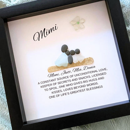 A personalized sea glass and pebble art piece with the inscription 'Mimi' in a shadowbox frame, accompanied by a floral decoration.