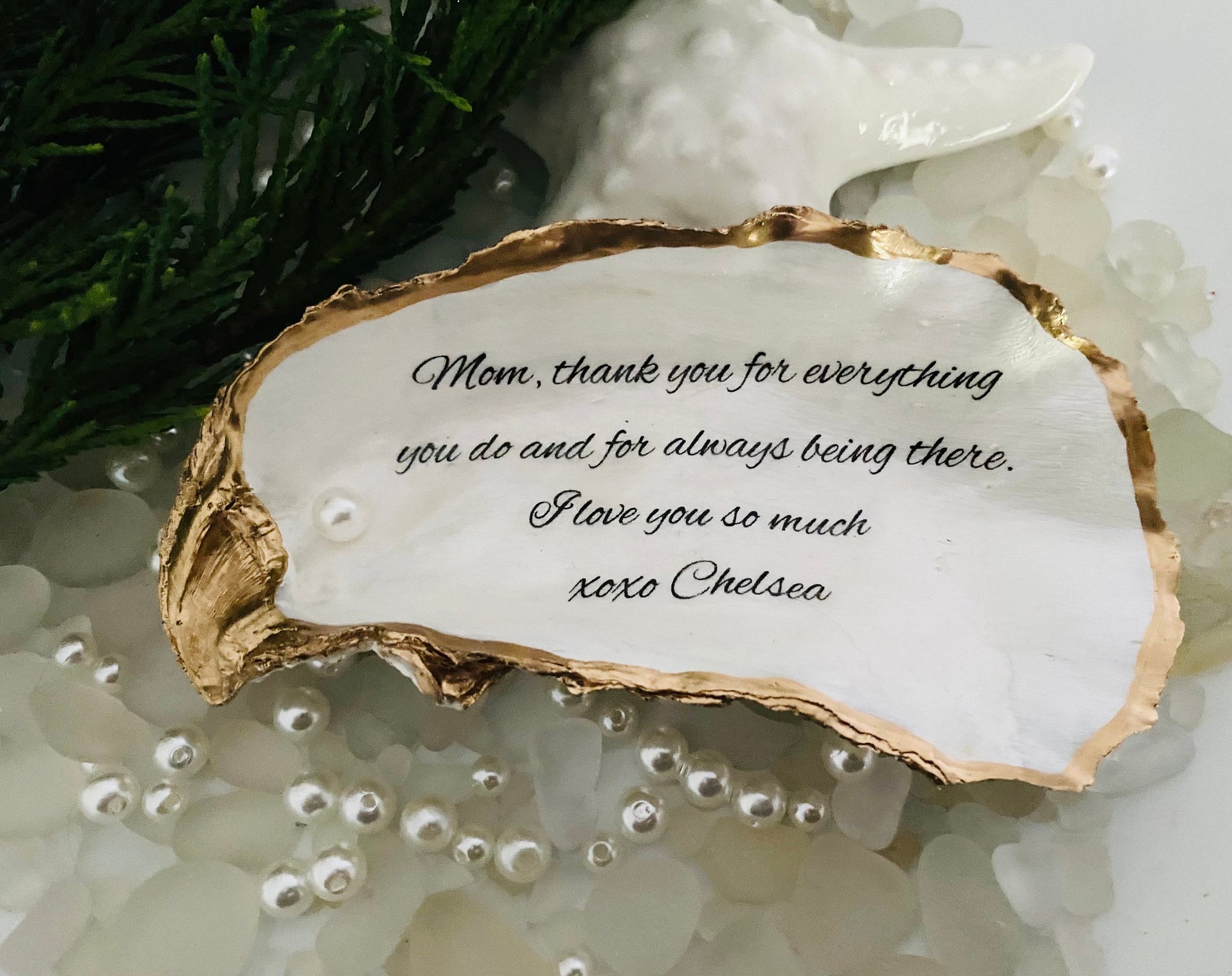 A personalized oyster shell jewelry dish with a message written on it, placed on a white surface surrounded by decorative pearls.