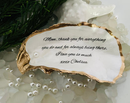 A personalized oyster shell jewelry dish with a message written on it, placed on a white surface surrounded by decorative pearls.