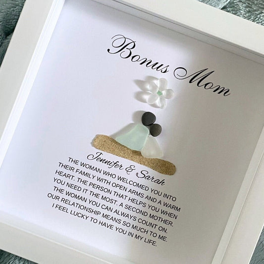 A personalized gift frame with a title 'BONUS MOM' at the top, featuring a sea glass figure and pebble art, placed on a soft surface.