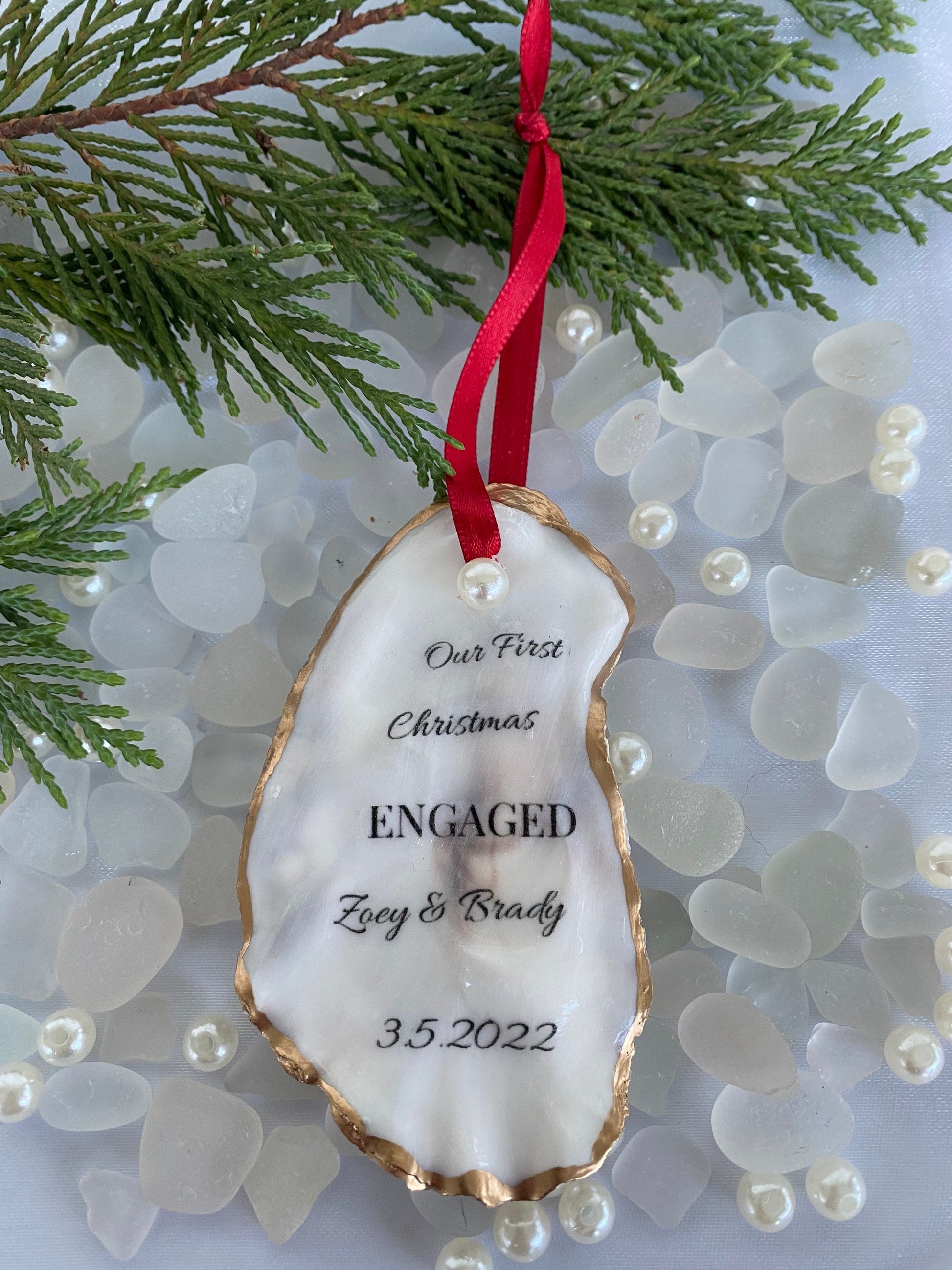 A personalized engagement ornament made of oyster shell with gold gilded edges, displaying a message that reads 'Our First Christmas Engaged', with a date '3.2.2022' below the text. The ornament is hanging from a red ribbon, placed against a backdrop of pine needles.