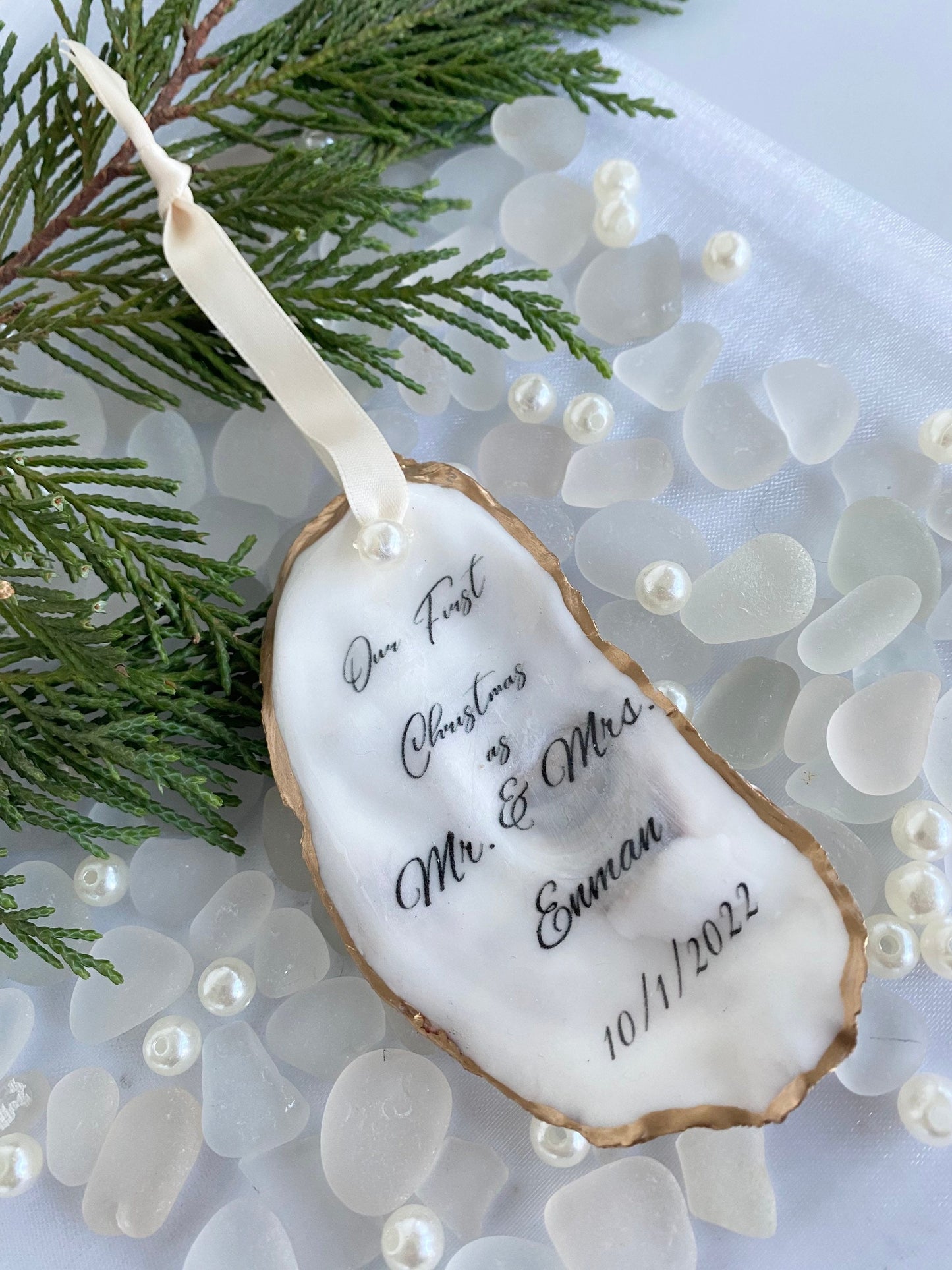 A personalized wedding Christmas ornament made of oyster shell with the names 'Mr & Mrs' and a date '11/27/2024'  on it, placed on a branch with pine needles and decorative pearls around.