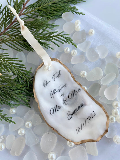 A personalized wedding Christmas ornament made of oyster shell with the names 'Mr & Mrs' and a date '11/27/2024'  on it, placed on a branch with pine needles and decorative pearls around.