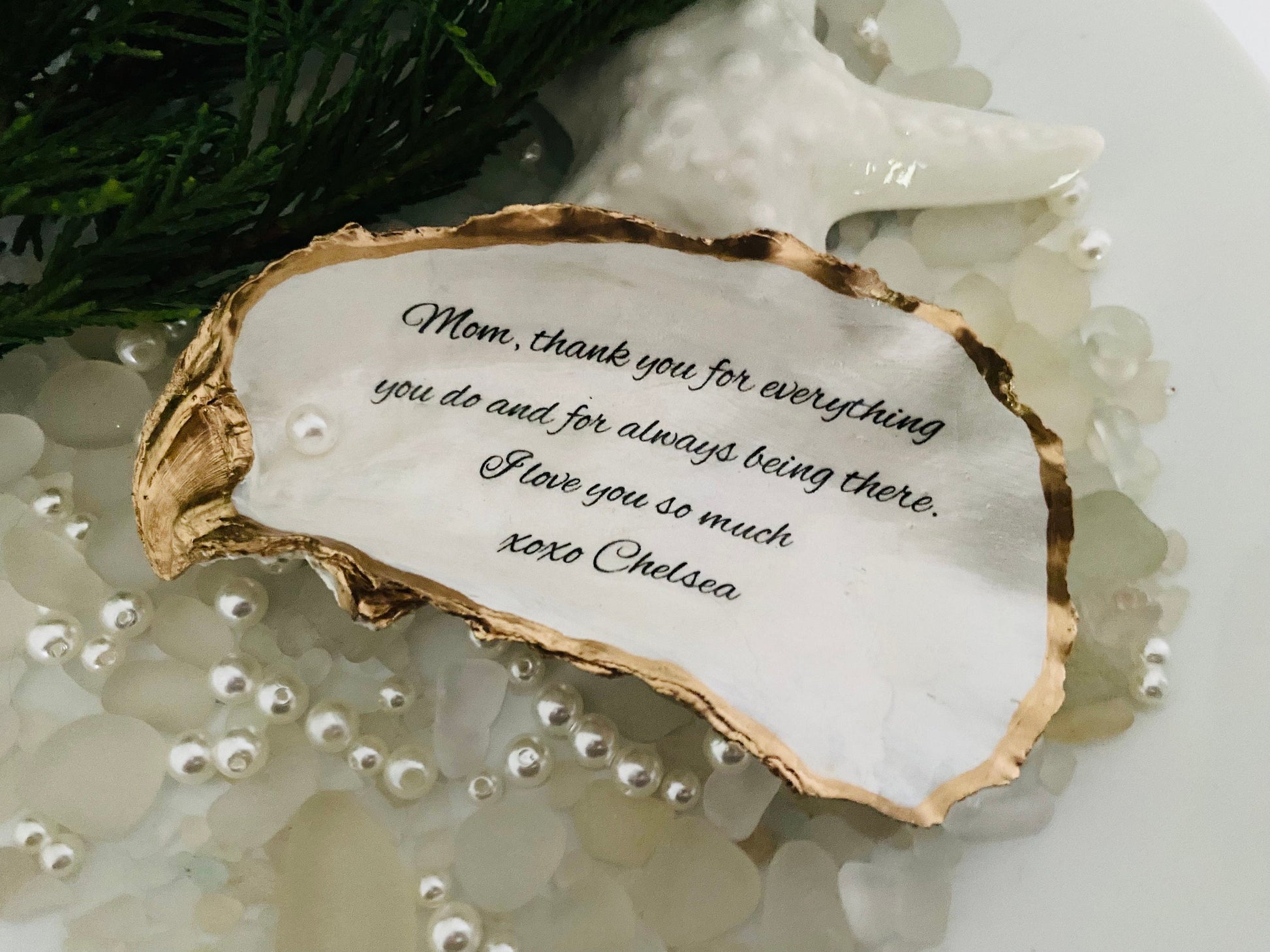 A personalized oyster shell jewelry dish with a message written on it, placed on a white surface surrounded by decorative pearls.