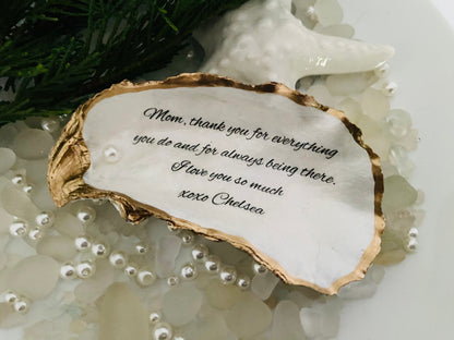 A personalized oyster shell jewelry dish with a message written on it, placed on a white surface surrounded by decorative pearls.