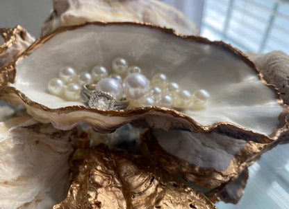 A decorative oyster shell ring dish with a glossy finish and gold leaf trim, displayed as a bridal party gift with pearl accents.
